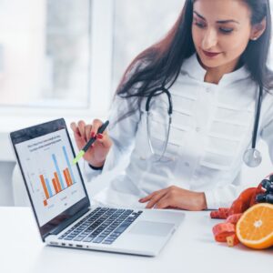 Female nutritionist in white coat sitting indoors in the office at workplace with laptop